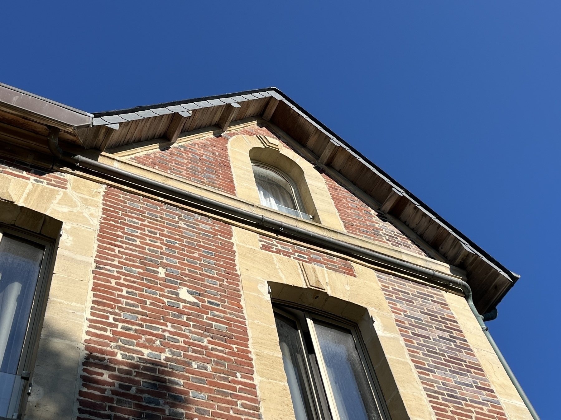 A wooden gabled roof looked at from the ground looking up. Pale yellow sandstone frames two tall windows, one square and one arched. The sunshine lights up the stonework and the red bricks. The sky is deep blue with no clouds above. 