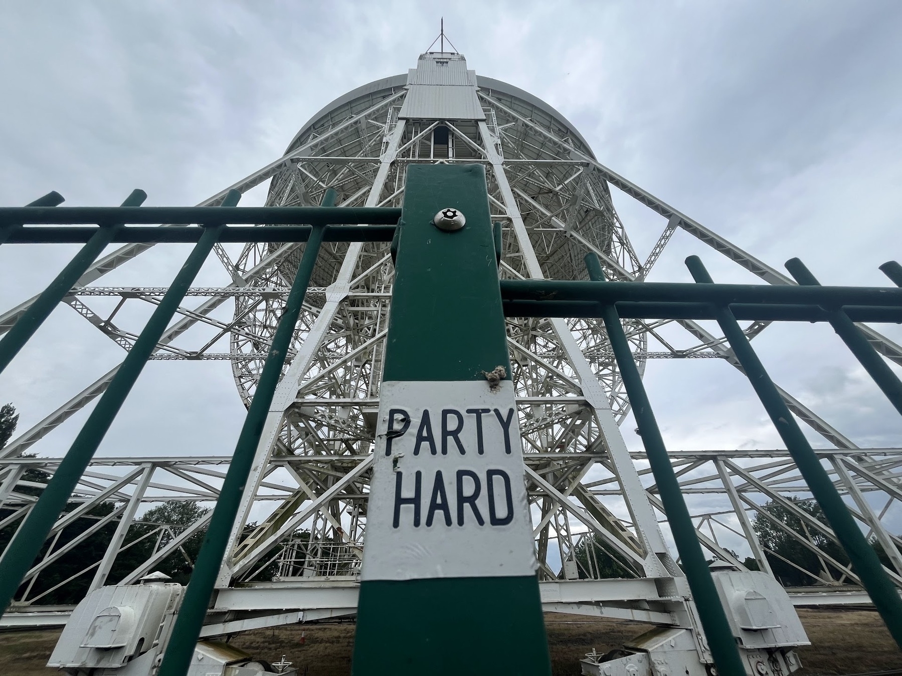 A giant radio telescope dish points up towards a cloudy grey sky. Stell girders painted white and stained with rust are seen supporting the dish, and in the foreground, a modern green painted metal fences with a small white sticker on it that says "party hard" in a simple black font on a white sticker.