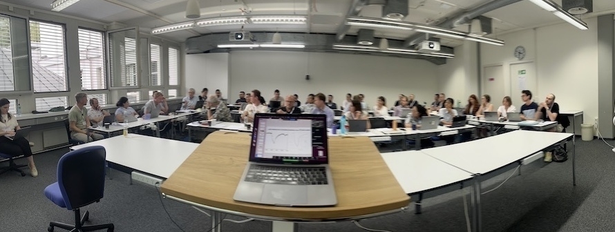 A panoramic photo of the crowd at the Exocomets meeting. A laptop sits on the lectern in the middle of the photo. The room is white with windows along the left wall.