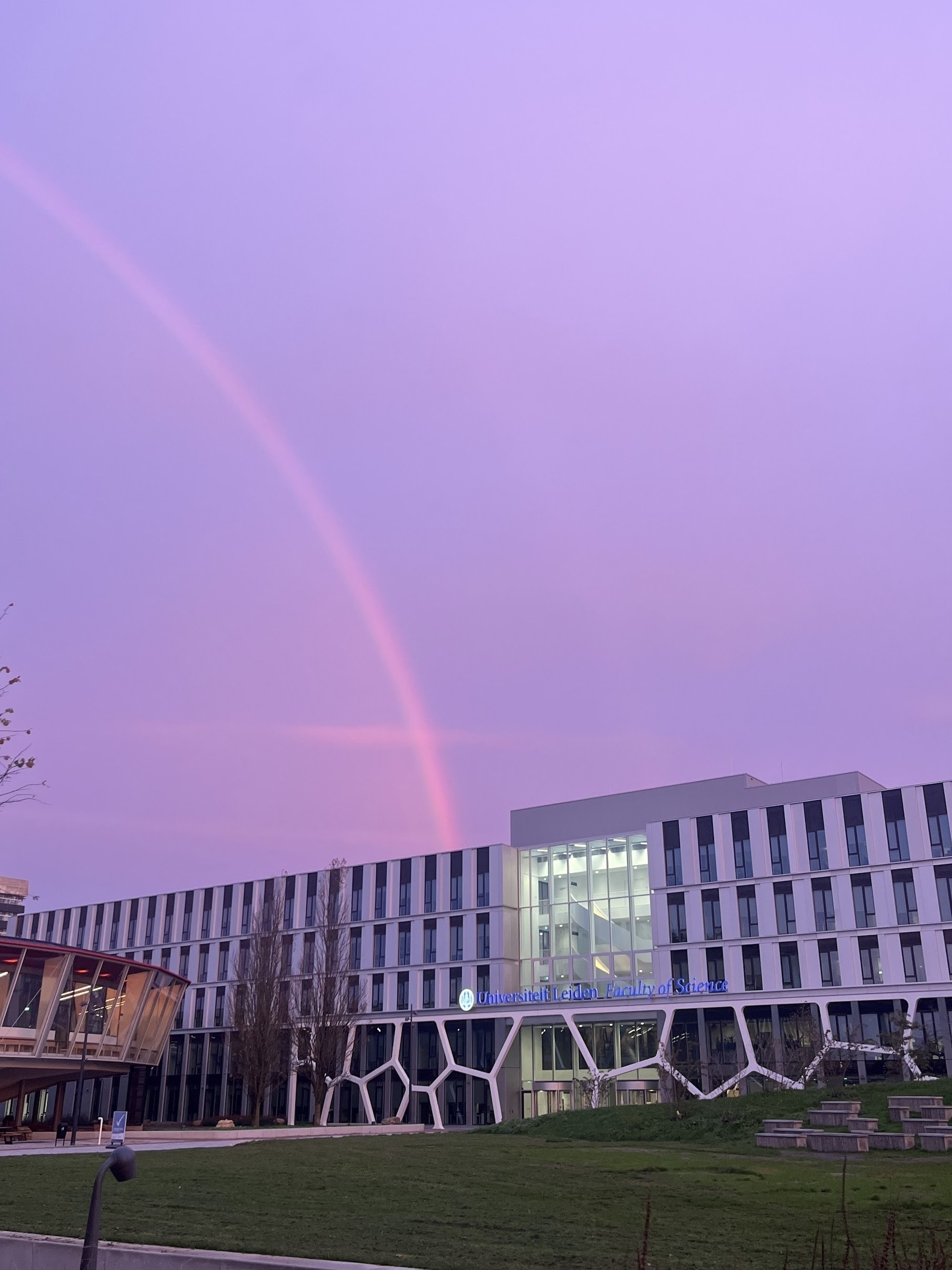 A purple sky with a rainbow generated just aster sunrise, with the rainbow touching a long white building with the blue sign "Leiden Univsrsity Faculty of Science". The rainbow terminates at the top floor where the Leiden Observatory is housed.