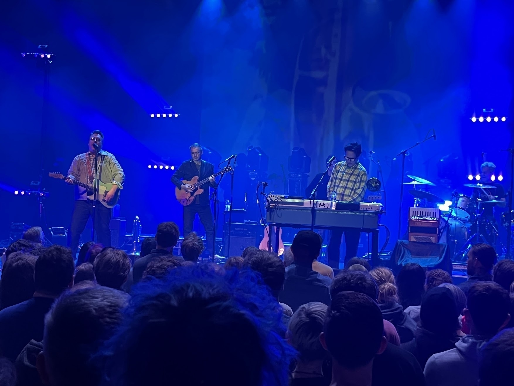 The two Johns, one in a plaid shirt, the other with a rocking guitar, on a blue lit stage with one othe band member. The heads of enthusiastic fans are along the bottom.