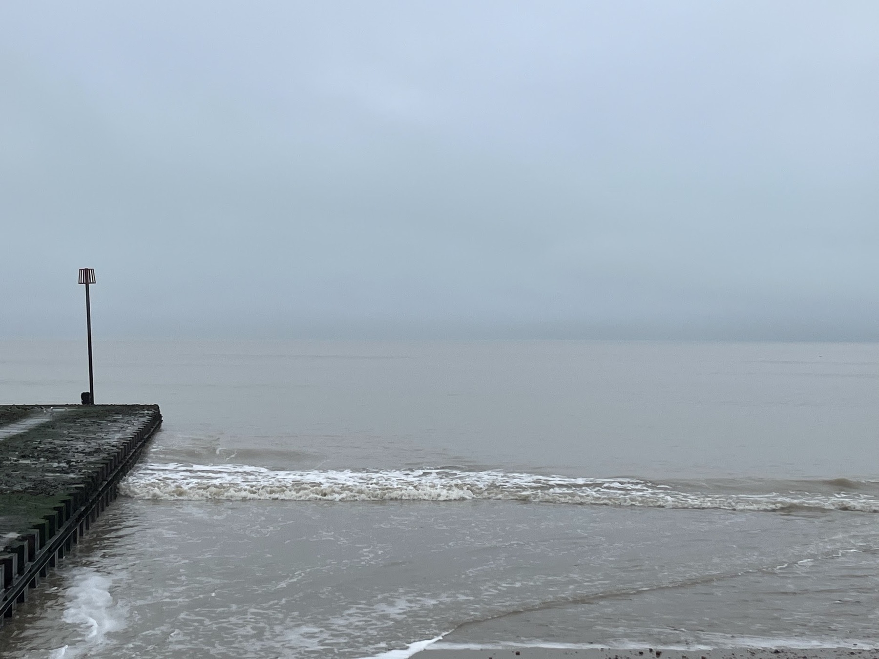 Gray featureless cloud down to the horizon, where it meets the gray sea. There is one sultry wave sloshing in reluctantly. ON the left is a concrete pier extending about 20 metres into the ocean, with a red metal basket on a pole indicating it's a rain outflow pipe. There is nothing else moving or colourful in the image.