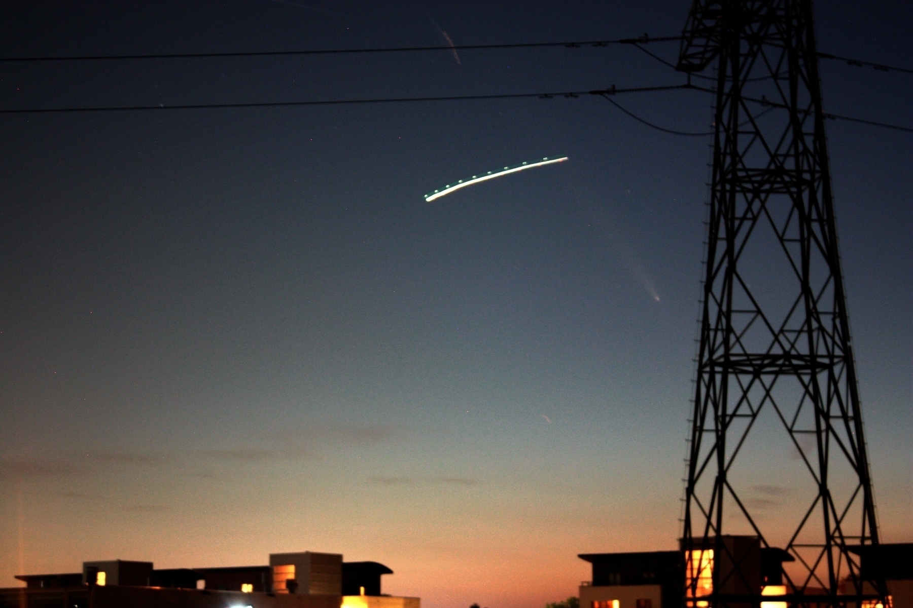 A twilight sky with the silhouettes of an electrical transmission tower and buildings. A smeared out plane trail can be seen, and the white head and fuzzy tail points upwards away from the direction of the setting sun. 