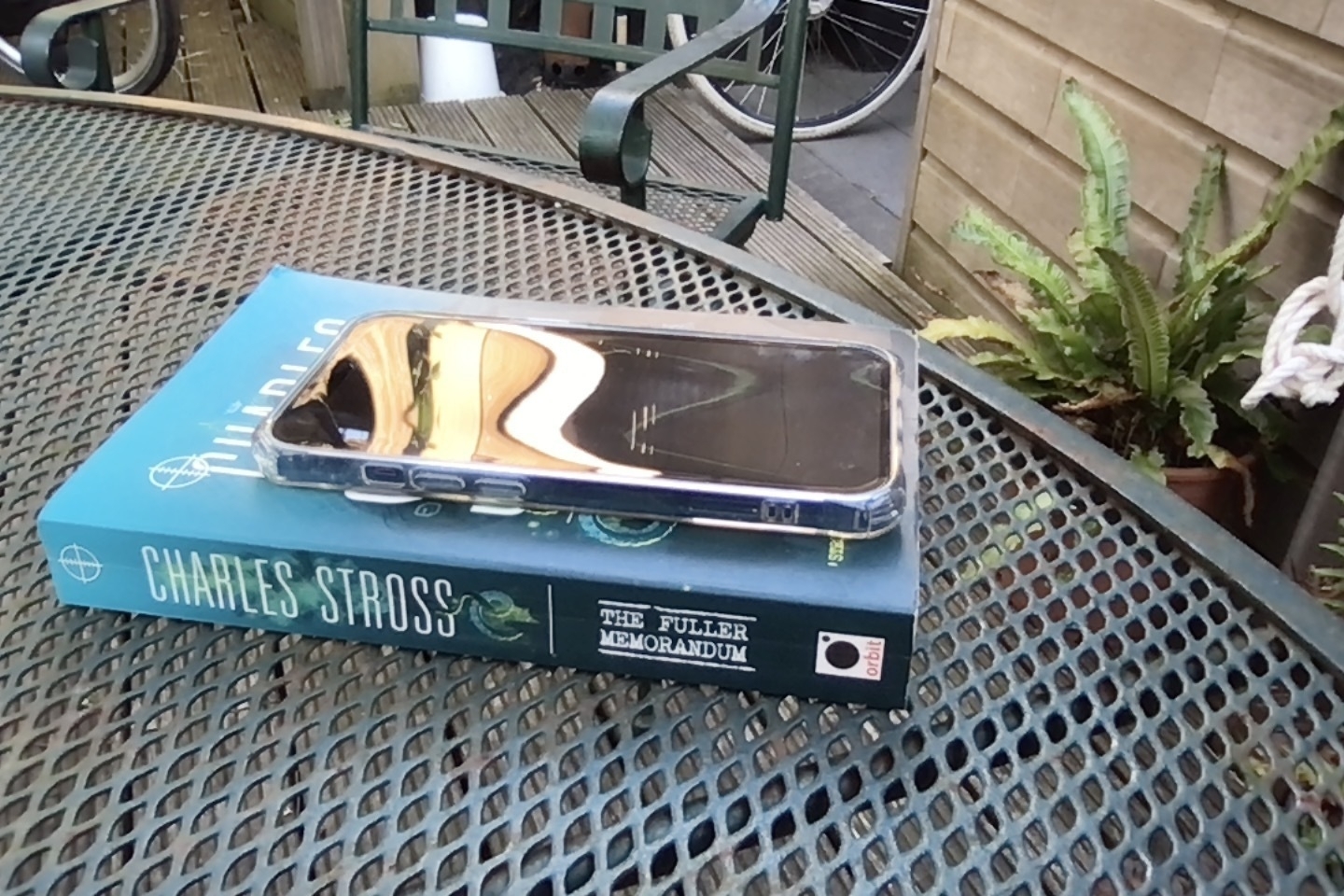 An iPhone sits on top of a copy of Charles Stross' The Fuller Memorandum, which itself is on a green garden table. The blue sky and part of a building are reflected in the phone screen, and the straight white lines of the building roof are distorted into a sinuous S shape in the reflection of the phone screen.