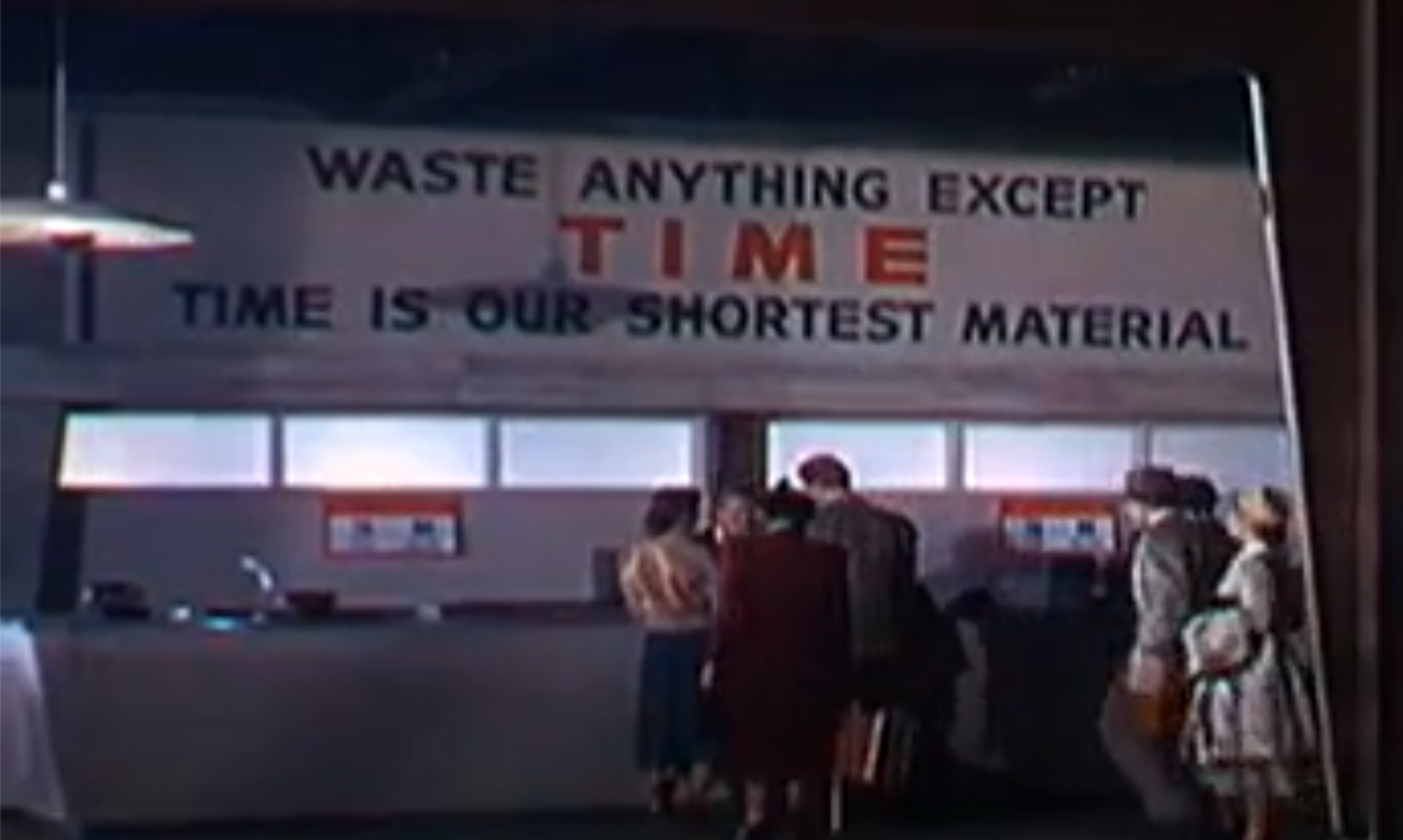 A sign that reads "Waste anything except time. Time is our shortest material." The sign is hanging inside a laboratory with men and women standing underneath it in 1950's clothing.