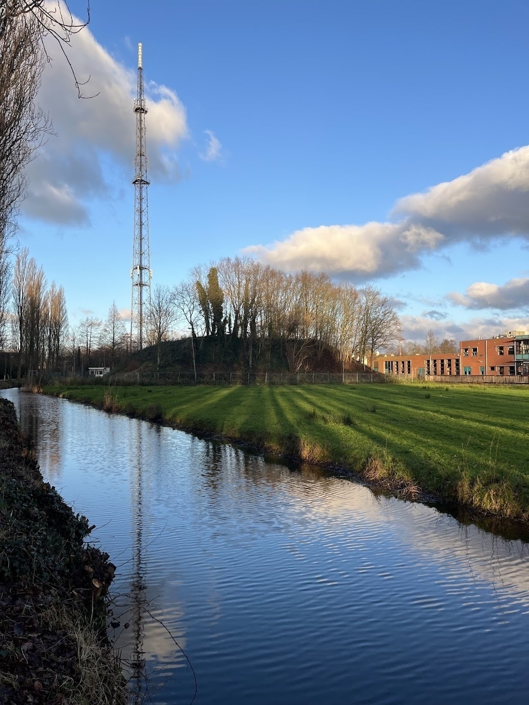 Blue skies over a large radio tower on the left, next to a low trapezoidal structure covered in winter trees. This is a concrete bunker built during the Cold War, covered in soil and several trees. In the foreground, a canal shows a reflection of the radio tower.