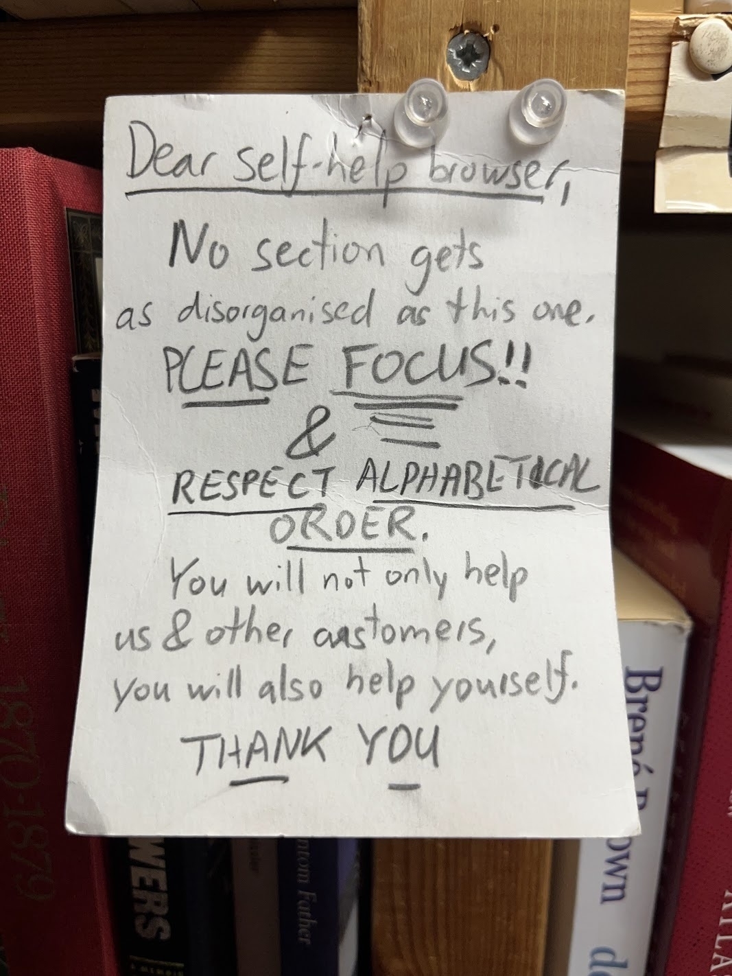 A paper card pinned up on a wooden shelf in a bookshop. The card has somewhat frantically written words in a pencil that may have been pressed a bit too hard, as if the author was extremely frustrated. The card reads: "Dear self-help browsers: no section gets as disorgansied as this one. PLEASE FOCUS! and respect alphabetical order. You will not only help us and the other customers, you will also help your self. THANK YOU."