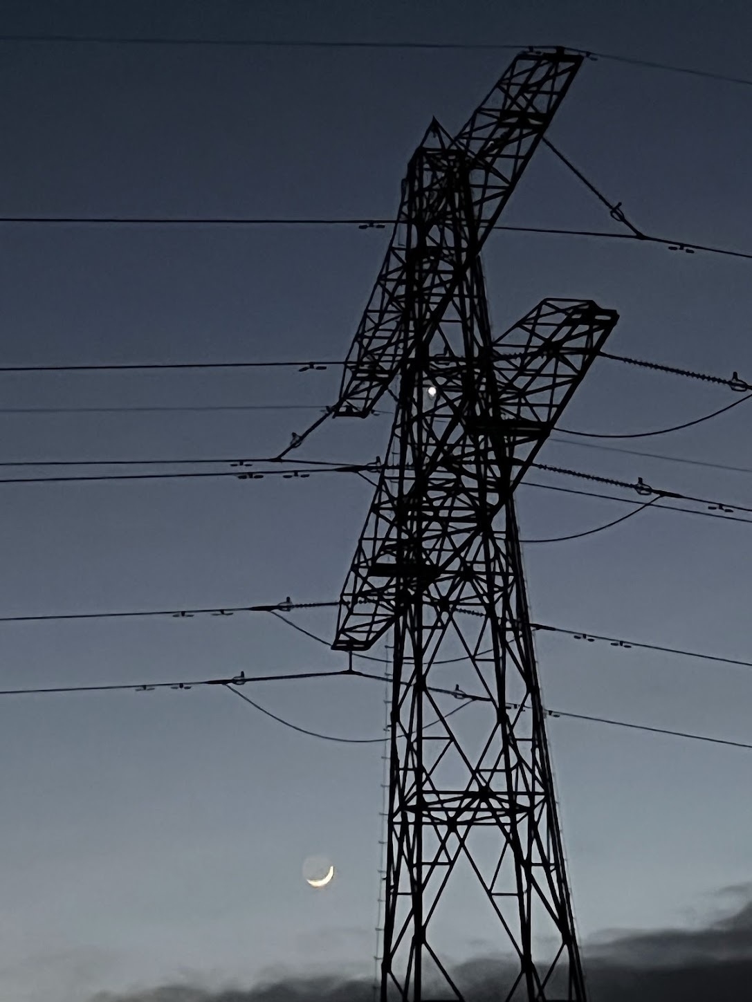 The deepening twilight sky with the silhouette of an electricity pylon in the foregroud, taken from a low angle looking up. The thin crescent Moon is in the lower part, with Earthshine visible. Venus glows brightly in between the pylon framework.