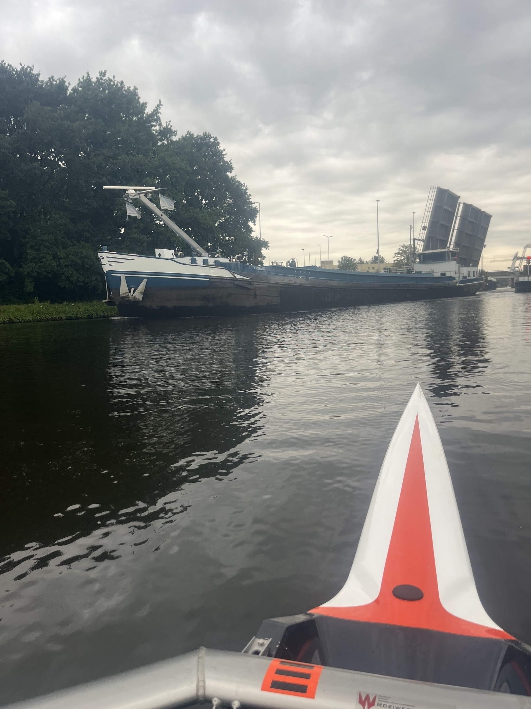 A grey sky with a road bridge that has opened up to allow a Big Flipping Boat to pass under it. The boat is about 50 metres long and is painted blue and white. The photo is taken from the point of view looking from a single rowing shell, with the bow of the boat in the lower right of the image.