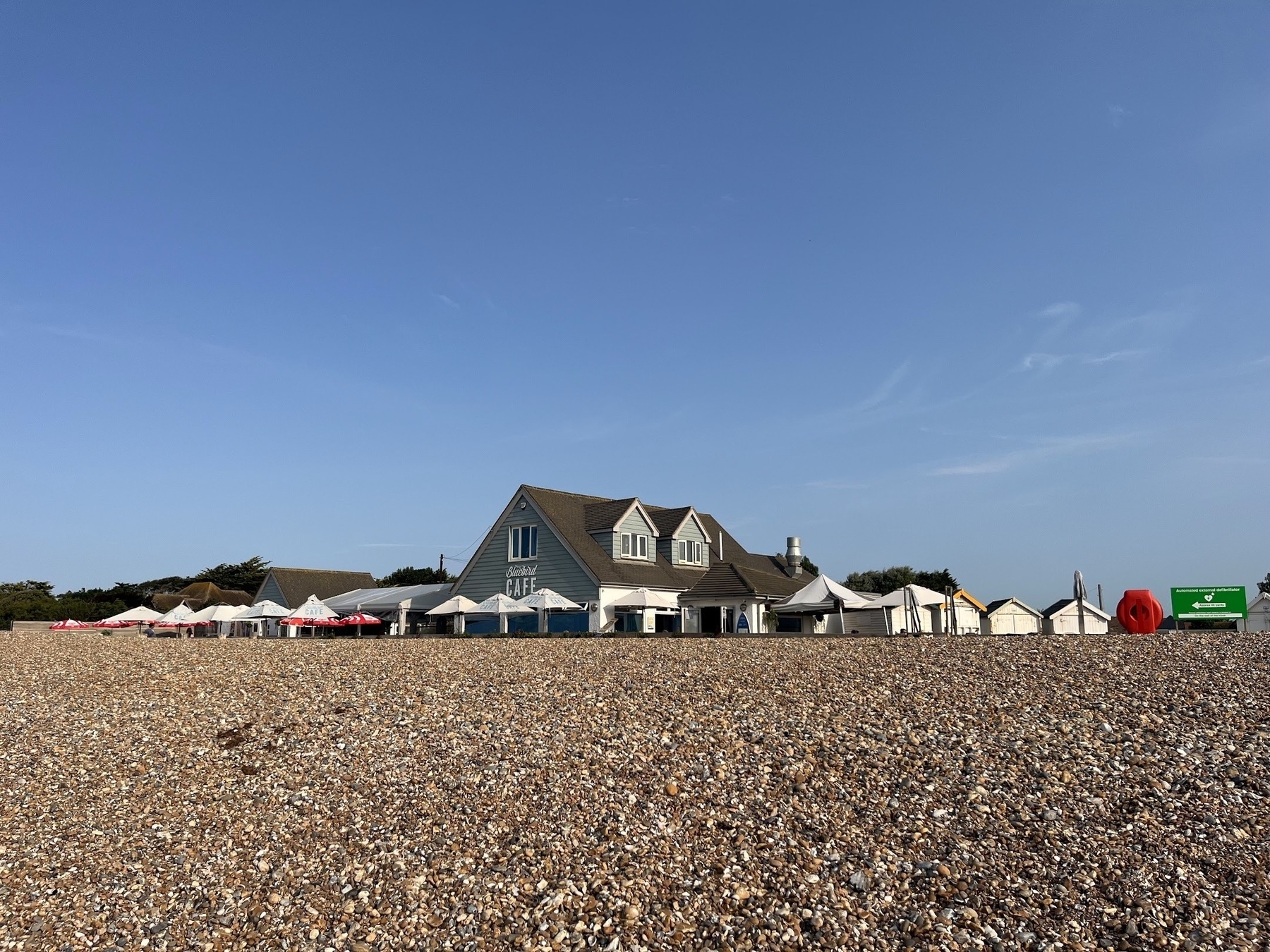 A pebble beach takes up the bottom half, a blue sky the top half. In the middle, a roofed two story building with dining tables around it, with "Bluebird Cafe" printed on the side of it.
