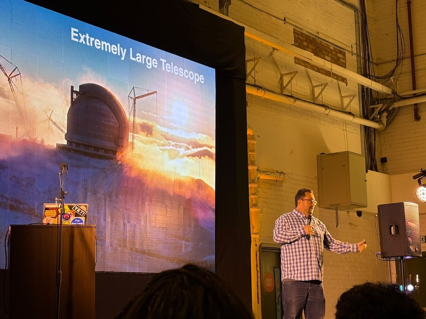 An image of the Extremely Large Telescope shrouded in mist with a rising sun is projected on a large screen, with me in a blue and white checked shirt standing in front of it with me waving my arms around excitedly and talking to the audience.