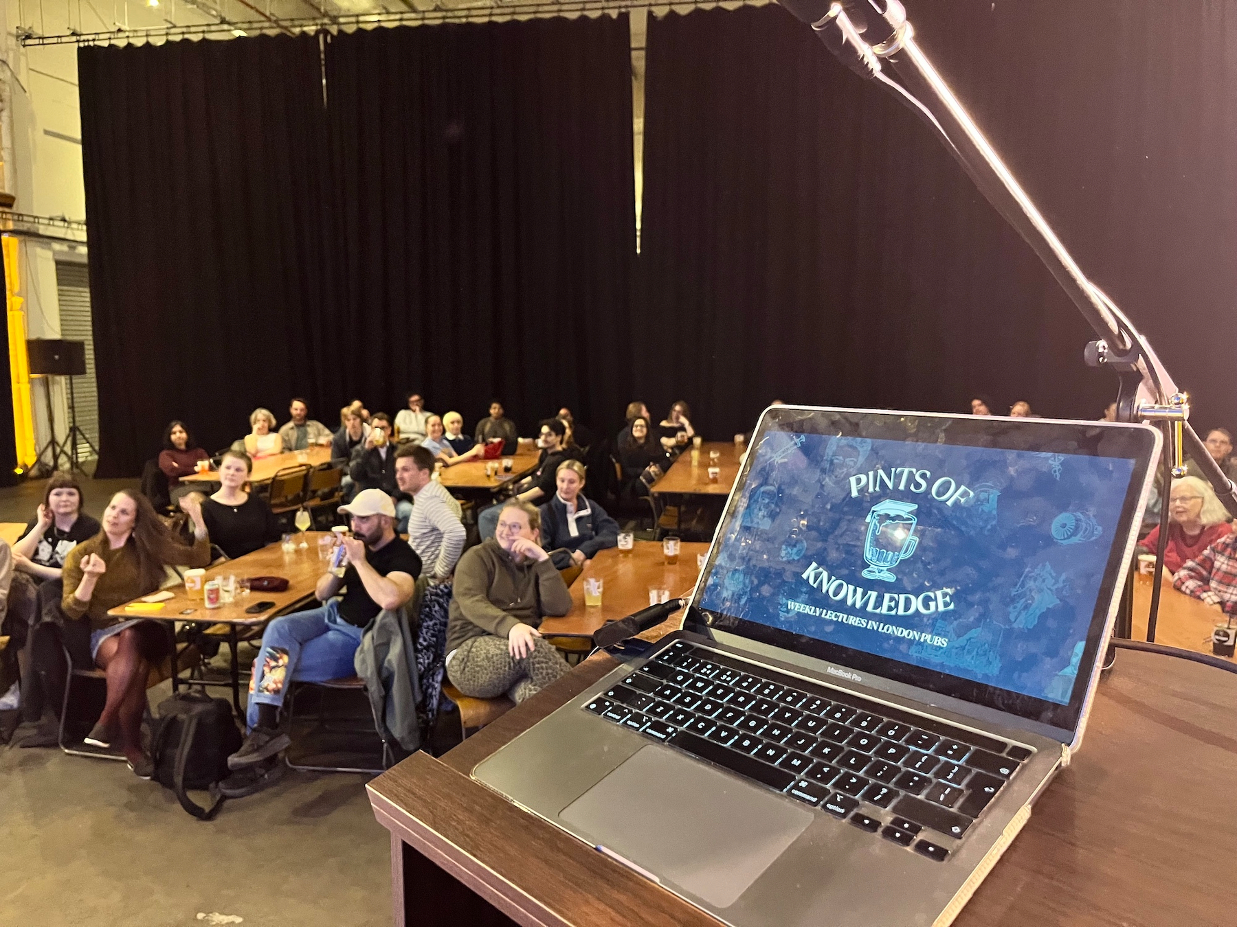 View from the stage with a fingerprint smudged laptop screen (not my laptop!) with “Pints of Knowledge” logo on it, and in the background several beer tables with people around them, looking at the compare talking off picture left. A large black stage curtain is at the back of the space.
