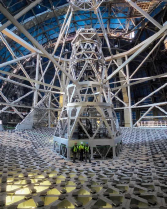 Three astronomers standing in the middle of a giant triangular array of mirror support pads which will hold the Extremely Large Telescope's primary mirror surface. The array is backlit to show the support girders.