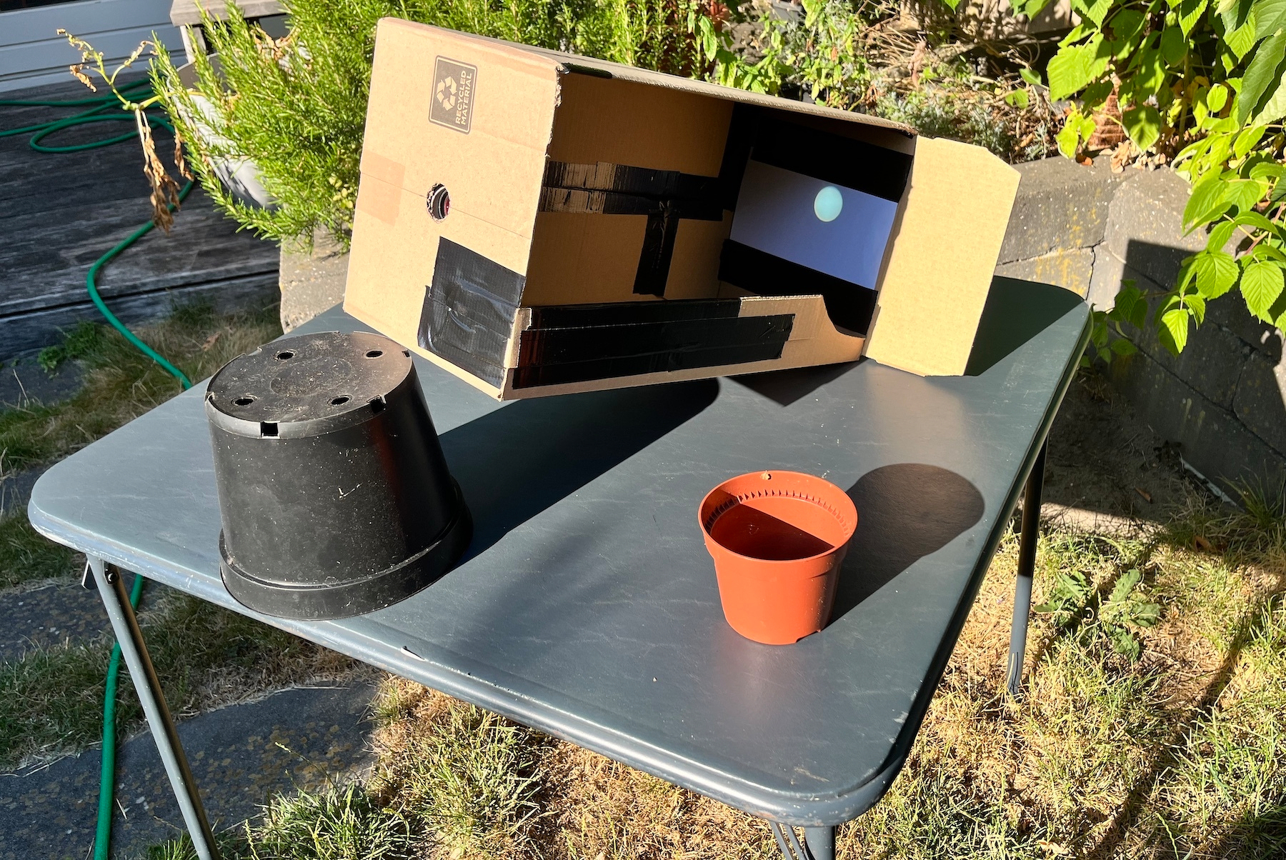 A cardboard box covered in duct tape, propped up by an upturned black plant pot, sitting on a cheap blue card table, itself standing in a garden with a brick wall and bushes behind it. The side of the cardboard box is missing and an image of the Sun can be seen projected on a sheet of white paper duct taped onto the inside back of the cardboard box.