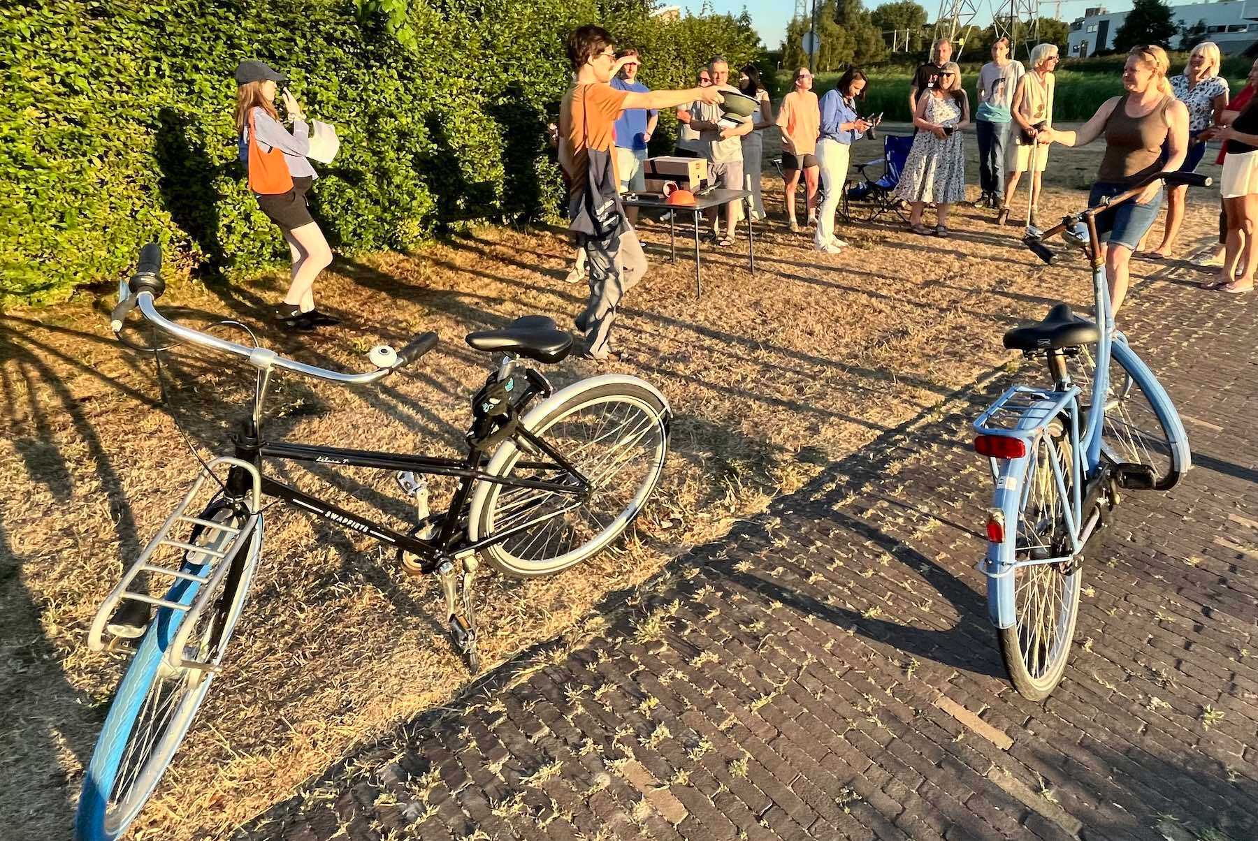 About a dozen people are standing in the setting sun and talking. They are standing around the card table with the solar projector sitting on a card table. One person is wearing solar glasses and is looking at the setting sun, around the time of maximum eclipse. There are two large bikes propped up on their kick stands in the foreground. 